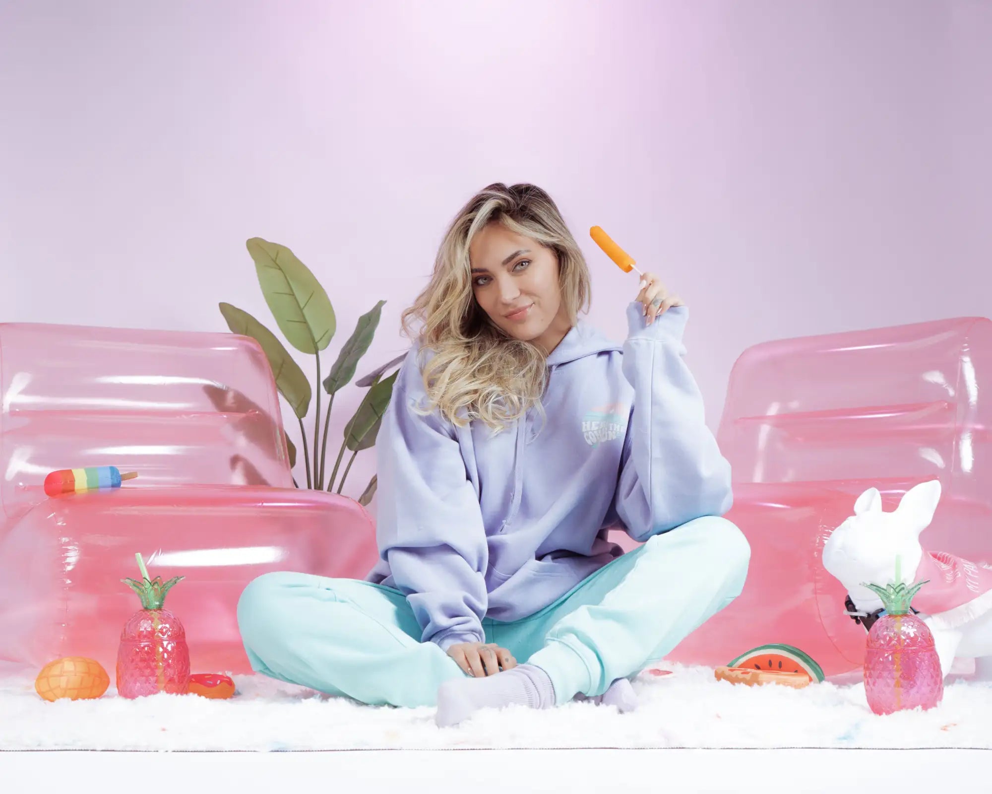 A soft lavender hoodie with a subtle rainbow logo on the chest, worn by a person sitting cross-legged.